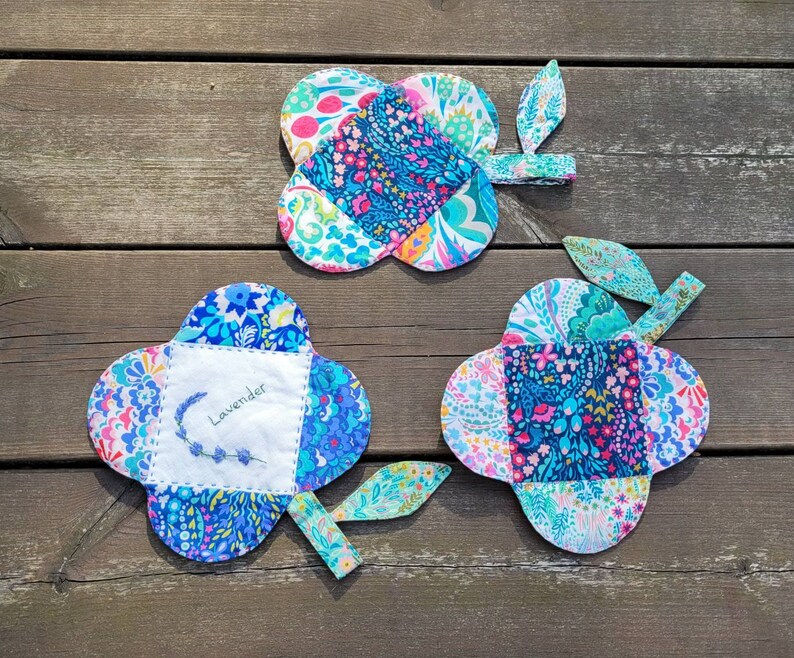 Pu&ograve; includere: Tre sottopentola in tessuto colorati a forma di quadrifoglio con un gambo. I sottopentola sono realizzati in tessuto patchwork con una variet&agrave; di motivi floreali in tonalit&agrave; di blu, rosa, verde e bianco. Un sottopentola ha la parola "Lavender" ricamata.