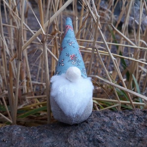 Pode incluir: Um gnomo de tecido azul e branco com barba branca e nariz de pompom branco. O gnomo está sentado em uma rocha com grama seca atrás.