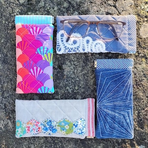 May include: Four quilted eyeglass cases in various designs. One case features a colorful fan pattern, another has a denim design, and a third has a floral pattern. The fourth case is denim with a white lace design. The cases are on a rock surface.