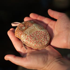 以下が含まれることがあります： ジッパー開閉とループが付いた、小さな柄の小銭入れ。茶色、ピンク、クリーム色の花柄と鳥のデザインが特徴です。小銭入れは開いた手の中にあります。