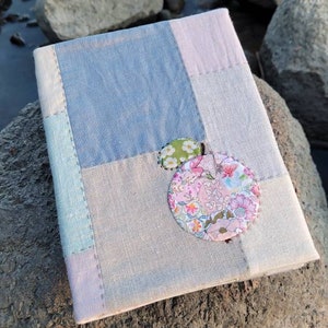 May include: A handmade journal with a patchwork cover in shades of blue, pink, and gray. The journal is adorned with two decorative fabric circles, one floral and one green with white polka dots. The journal rests on a large rock.