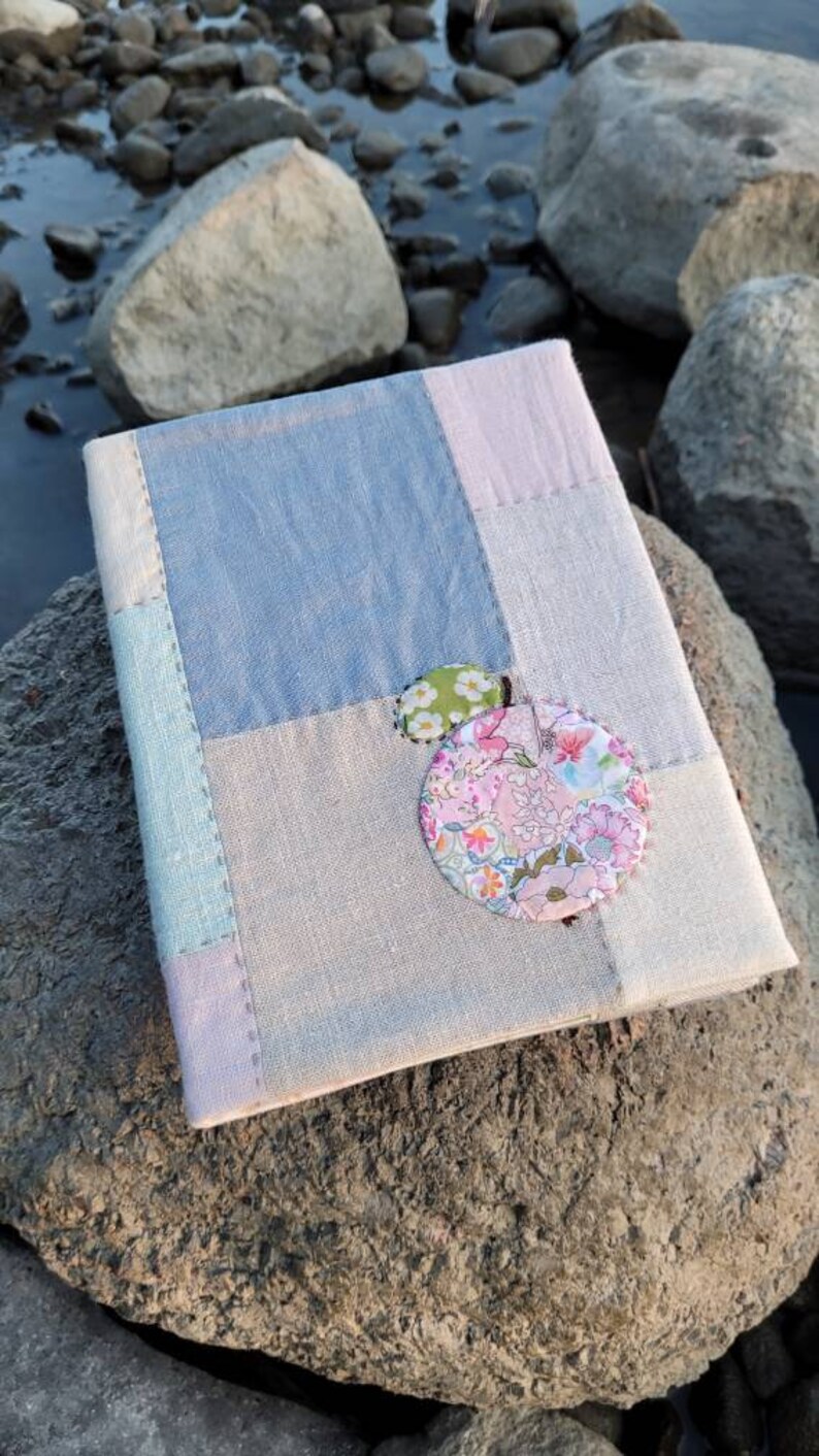 May include: A handmade journal with a patchwork cover in shades of blue, pink, and gray. The journal is adorned with two decorative fabric circles, one floral and one green with white polka dots. The journal rests on a large rock.
