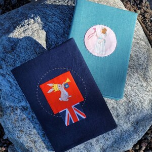 May include: Two fabric-covered books. One is teal with a circular patch featuring a girl holding a heart. The other is navy blue with a patch of a rabbit wearing a crown and blowing a horn, with a Union Jack design.