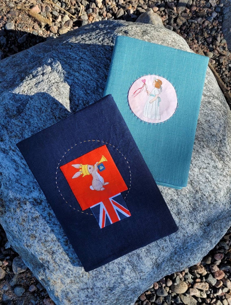May include: Two fabric-covered books. One is teal with a circular patch featuring a girl holding a heart. The other is navy blue with a patch of a rabbit wearing a crown and blowing a horn, with a Union Jack design.