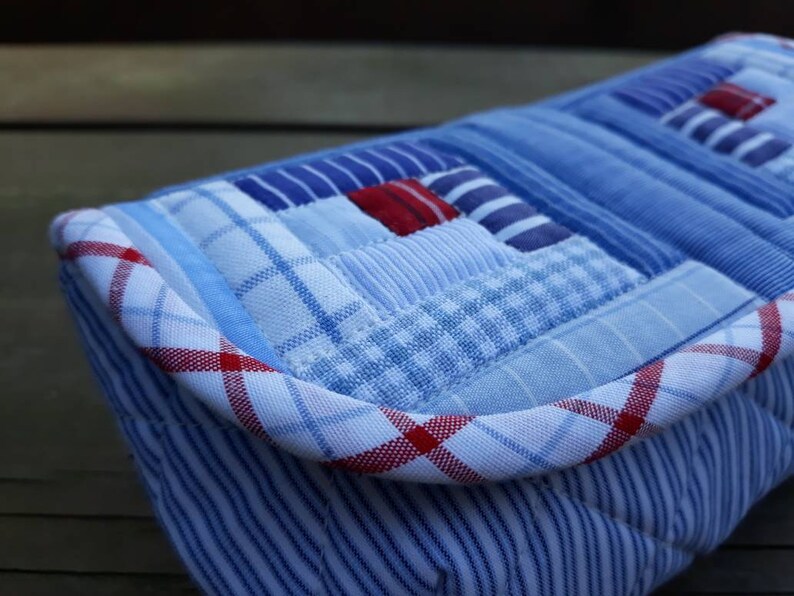 May include: A blue and white patchwork fabric clutch with a red and white plaid trim. The clutch is made from repurposed fabric and has a unique, handmade look.