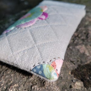 May include: A rectangular, quilted fabric pouch in a light gray, textured material. The pouch features a diamond-stitched pattern and is accented with colorful, patterned fabric patches in shades of pink, blue, green, and yellow at the corners.