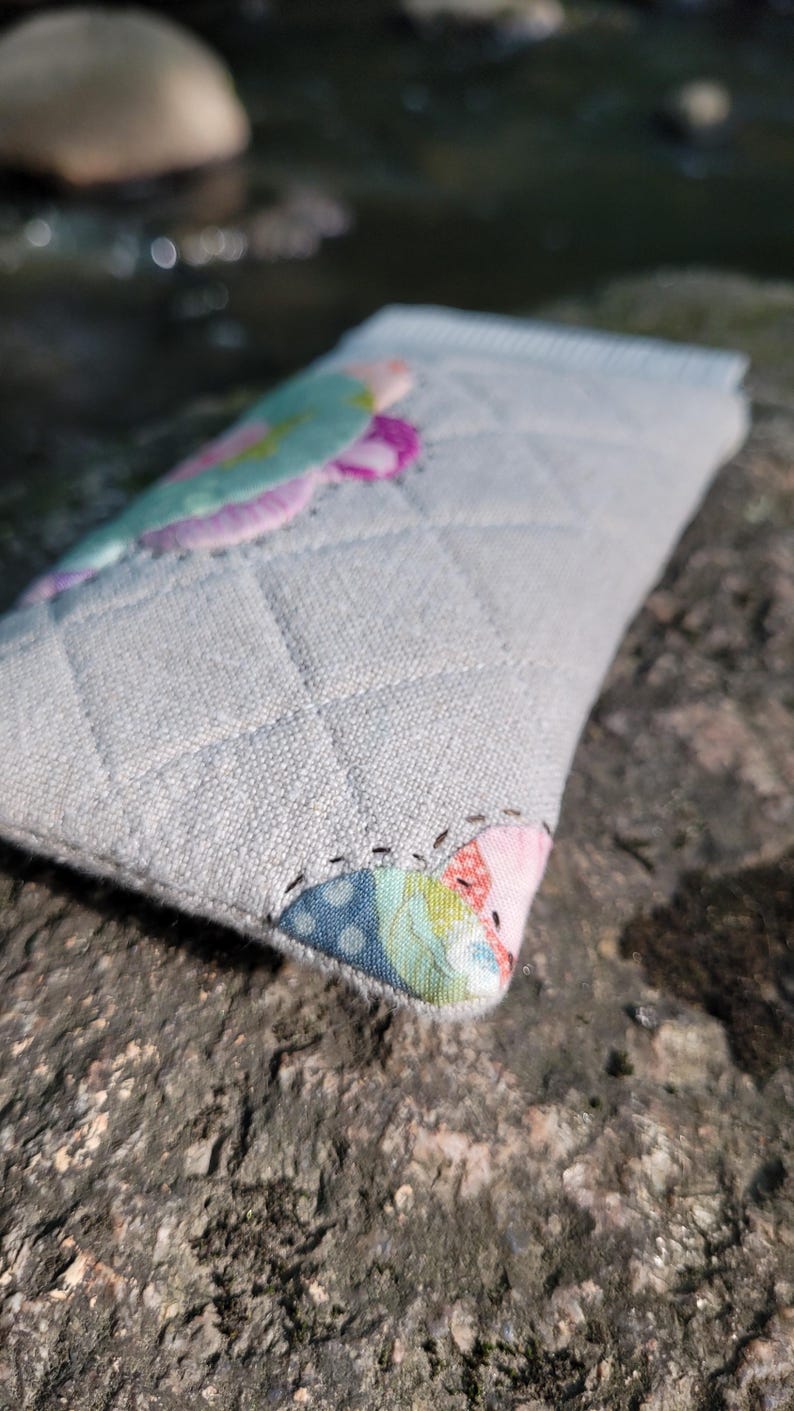 May include: A rectangular, quilted fabric pouch in a light gray, textured material. The pouch features a diamond-stitched pattern and is accented with colorful, patterned fabric patches in shades of pink, blue, green, and yellow at the corners.