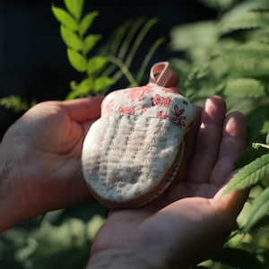 Puede incluir: Un pequeño objeto hecho a mano con forma de bellota, con un cuerpo beige claro y una parte superior con estampado floral en rojo y beige. El objeto tiene un diseño cosido y un pequeño lazo. El fondo es follaje verde borroso.