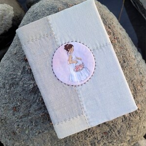 May include: A light gray linen journal or notebook with a stitched border. A circular patch on the cover features an illustration of a pregnant person in a white dress holding flowers. The journal rests on a gray rock.