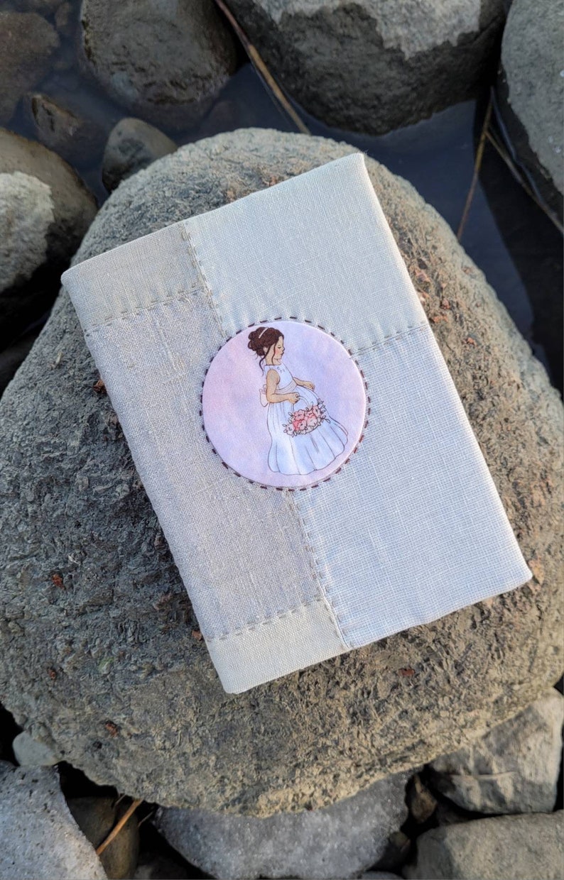 May include: A light gray linen journal or notebook with a stitched border. A circular patch on the cover features an illustration of a pregnant person in a white dress holding flowers. The journal rests on a gray rock.