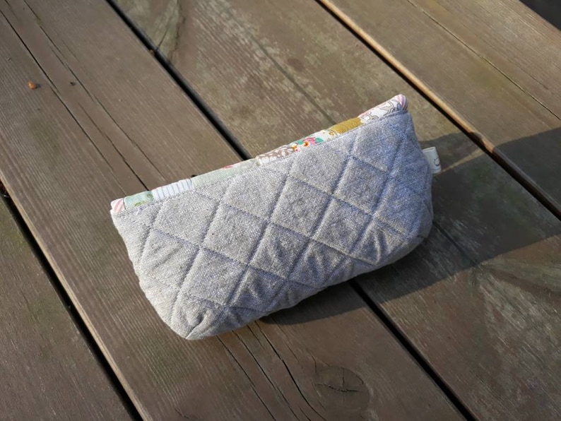 May include: A gray quilted zippered pouch with a floral patterned fabric lining. The pouch is lying on a wooden surface.