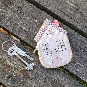 May include: A white quilted house-shaped keychain with red and white checkered fabric accents. The keychain has a small red heart on the front and a silver key attached to it.