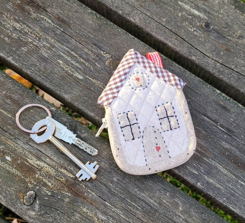 May include: A white quilted house-shaped keychain with red and white checkered fabric accents. The keychain has a small red heart on the front and a silver key attached to it.