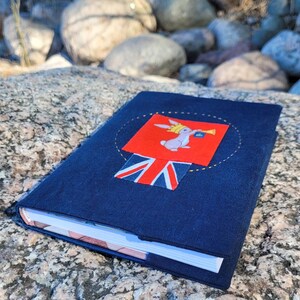 May include: A navy blue journal with a decorative patch featuring a rabbit wearing a crown and holding a trumpet, set against a red background. The patch includes a Union Jack flag. The journal is resting on a textured rock surface.