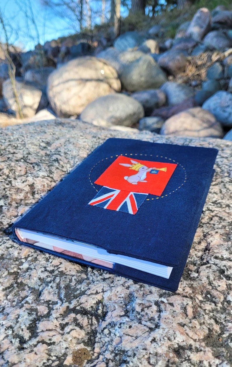 May include: A navy blue journal with a decorative patch featuring a rabbit wearing a crown and holding a trumpet, set against a red background. The patch includes a Union Jack flag. The journal is resting on a textured rock surface.