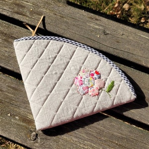 Puede incluir: Un calientavasos de tela acolchada beige con ribete a cuadros blanco y negro y una aplicación de flores de colores.