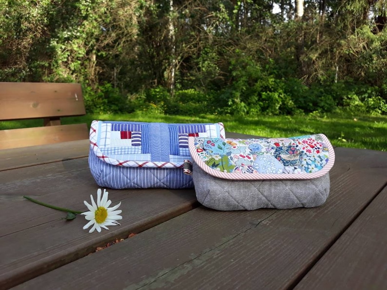 May include: Two fabric pouches, one with a blue and white patchwork design and the other with a floral print, are sitting on a wooden table.