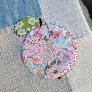 May include: Close-up of a patchwork design featuring a large, circular floral applique in shades of pink, green, and orange. The applique is stitched onto a light blue and beige fabric, with visible seams and a small green leaf detail.