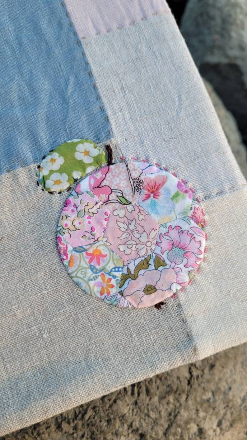 May include: Close-up of a patchwork design featuring a large, circular floral applique in shades of pink, green, and orange. The applique is stitched onto a light blue and beige fabric, with visible seams and a small green leaf detail.