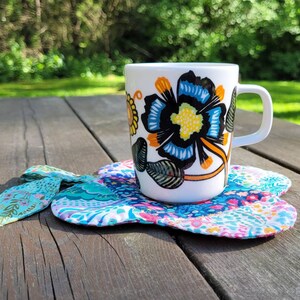 Pu&ograve; includere: Una tazza in ceramica bianca con un motivo floreale in tonalit&agrave; blu, gialle e nere. La tazza &egrave; appoggiata su un sottobicchiere di stoffa colorato.