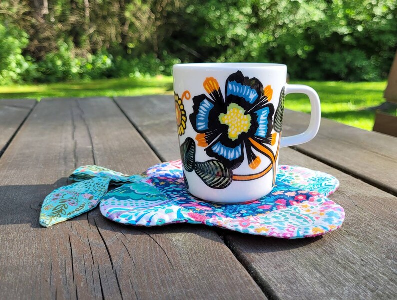 Pu&ograve; includere: Una tazza in ceramica bianca con un motivo floreale in tonalit&agrave; blu, gialle e nere. La tazza &egrave; appoggiata su un sottobicchiere di stoffa colorato.