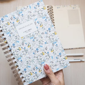 May include: A white spiral bound notebook with a floral pattern in shades of blue, yellow, and green. The cover features the text "DREAM. PLAN. DO." and "CARA ROSE Design Studio".