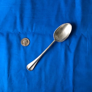 May include: A silver spoon with a rounded bowl and a long, slender handle. The spoon is lying on a blue fabric surface.