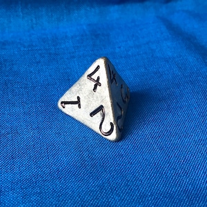 Puede incluir: Un dado de cuatro caras plateado con los números 1, 2, 3 y 4 en sus caras. El dado está sobre una superficie de tela azul.