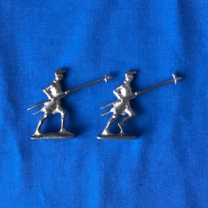 May include: Two silver toy soldiers in a running pose, each holding a spear with a cross-shaped tip. The figures are on a blue background. The soldiers are wearing hats and appear to be in motion.