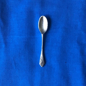 Puede incluir: Una cucharadita de plata con un cuenco redondeado y un mango puntiagudo. La cuchara está sobre un fondo de tela azul.