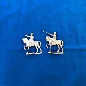 May include: Two silver figurines of a person on horseback, set against a blue backdrop. One figure points, the other holds a sword. The figures are detailed, showing the rider's attire and the horse's form. The theme suggests historical or military interest.