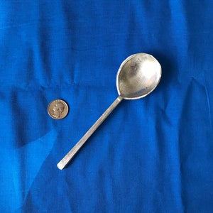May include: A silver spoon with a rounded bowl and a long, narrow handle. The spoon is lying on a blue fabric surface.