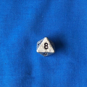 May include: A silver-colored, multi-sided die with the number 8 visible. The die is set against a blue fabric background. The die appears to be made of metal and has a worn, vintage look.