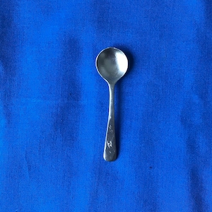 May include: A silver teaspoon with a rounded bowl and a long, slender handle. The spoon is sitting on a blue fabric surface.