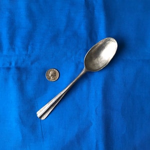 May include: A silver spoon with a rounded bowl and a long, slender handle. The spoon is lying on a blue fabric surface.