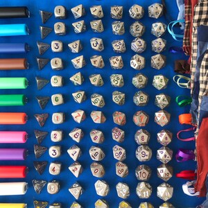 May include: A collection of metallic dice with various numbers and shapes, including D4, D6, D8, D10, D12, and D20, arranged on a blue surface. Also includes a US quarter and a set of colorful pens.