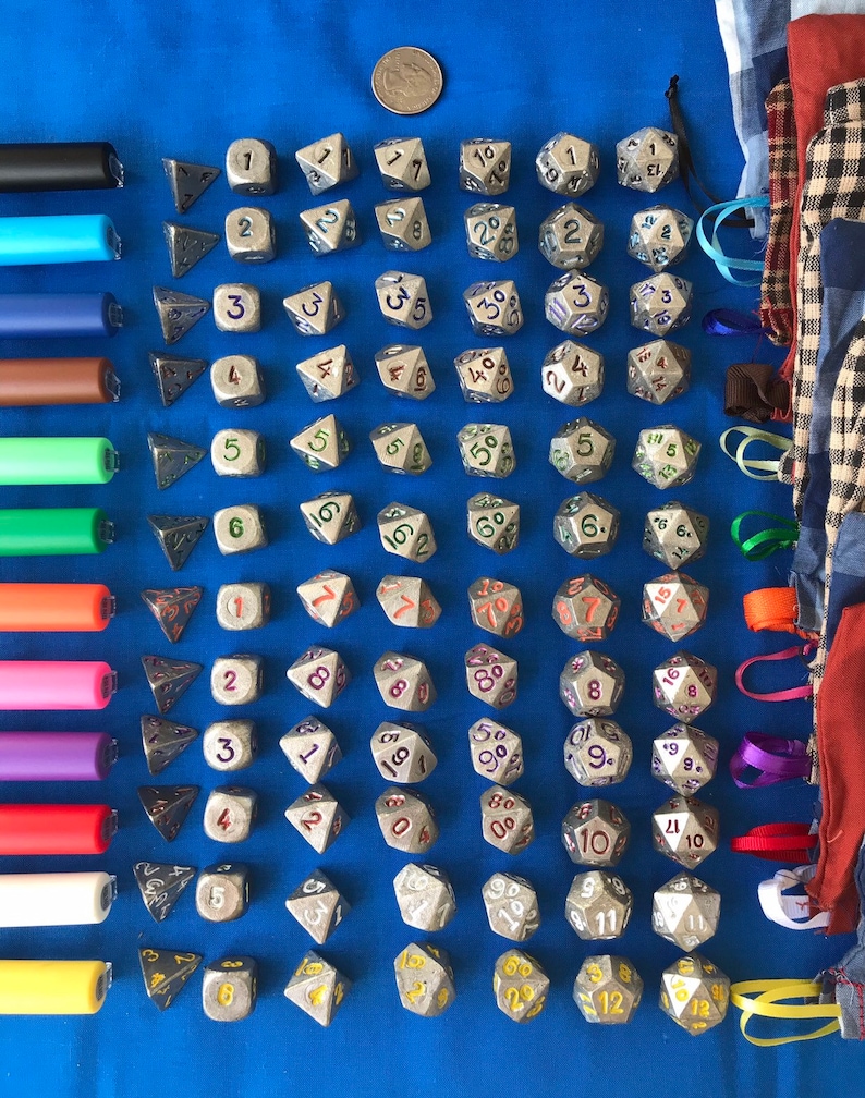 May include: A collection of metallic dice with various numbers and shapes, including D4, D6, D8, D10, D12, and D20, arranged on a blue surface. Also includes a US quarter and a set of colorful pens.