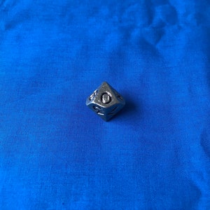 Peut inclure: Un dé en métal argenté avec des chiffres noirs. Le dé affiche le numéro 1.