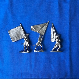 Puede incluir: Tres figuras en miniatura de soldados de estaño sosteniendo banderas. Las banderas son blancas y tienen un acabado plateado. Los soldados llevan uniformes plateados y están de pie sobre una superficie azul.
