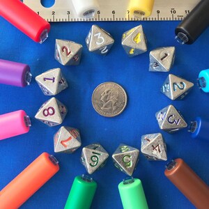 May include: A collection of silver polyhedral dice with numbers in various colors, arranged around a quarter coin on a blue surface. Colorful pens in red, purple, pink, orange, green, brown, blue, yellow, white, and black surround the dice. A metal ruler is also present.