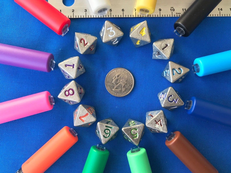 May include: A collection of silver polyhedral dice with numbers in various colors, arranged around a quarter coin on a blue surface. Colorful pens in red, purple, pink, orange, green, brown, blue, yellow, white, and black surround the dice. A metal ruler is also present.