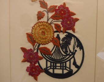 May include: Embroidered artwork featuring a floral design with orange, red, and pink flowers, and leaves. A circular black silhouette of a pagoda is also included in the design. The artwork is framed in a white mat.