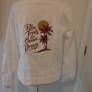 Może przedstawiać: Biała kurtka dżinsowa z bordową grafiką przedstawiającą palmy, słońce i napis "Palm Trees & Ocean Breeze" na plecach. Posiada zapięcia na guziki i kieszenie.