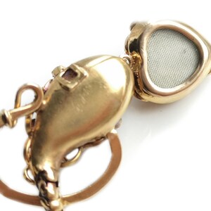May include: A gold heart-shaped locket with a hinged lid and a small, round, mesh-like window on the front. The locket is attached to a gold chain with a decorative clasp.