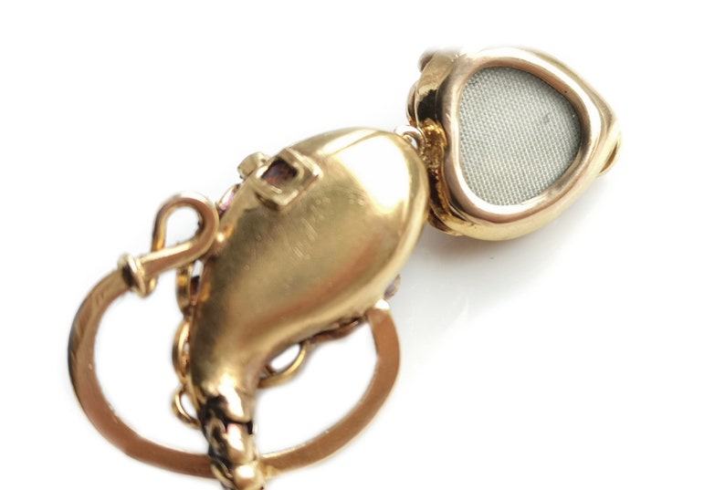 May include: A gold heart-shaped locket with a hinged lid and a small, round, mesh-like window on the front. The locket is attached to a gold chain with a decorative clasp.