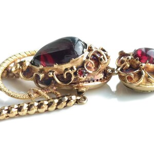 May include: A gold necklace with a red gemstone pendant. The pendant is ornate and features intricate details. The necklace is made of a chain with a twisted design.