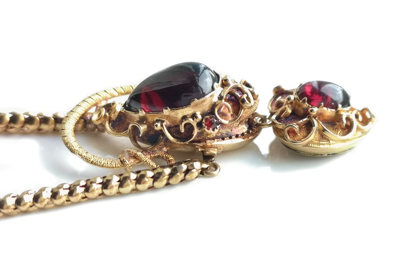 May include: A gold necklace with a red gemstone pendant. The pendant is ornate and features intricate details. The necklace is made of a chain with a twisted design.