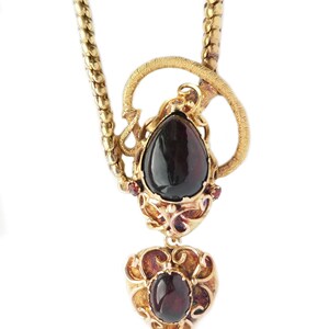May include: A gold necklace with a pendant featuring two dark red gemstones set in intricate gold filigree. The pendant is shaped like a heart with a teardrop-shaped gemstone above it.