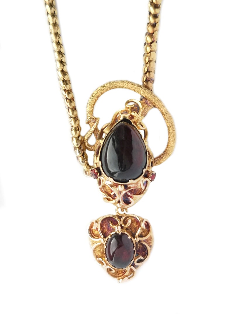 May include: A gold necklace with a pendant featuring two dark red gemstones set in intricate gold filigree. The pendant is shaped like a heart with a teardrop-shaped gemstone above it.