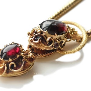 May include: A gold necklace with a pendant featuring two red gemstones set in ornate gold frames. The pendant is attached to a delicate gold chain.